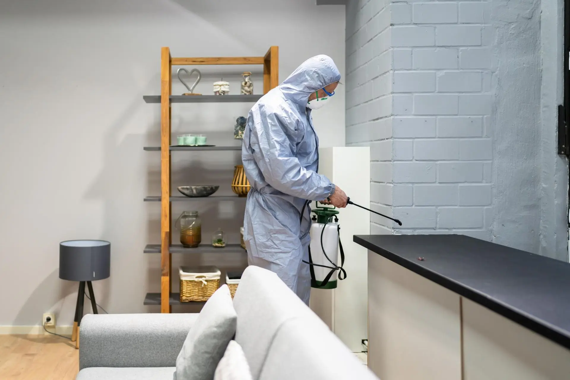 A man in a protective suit is spraying a living room with a sprayer.